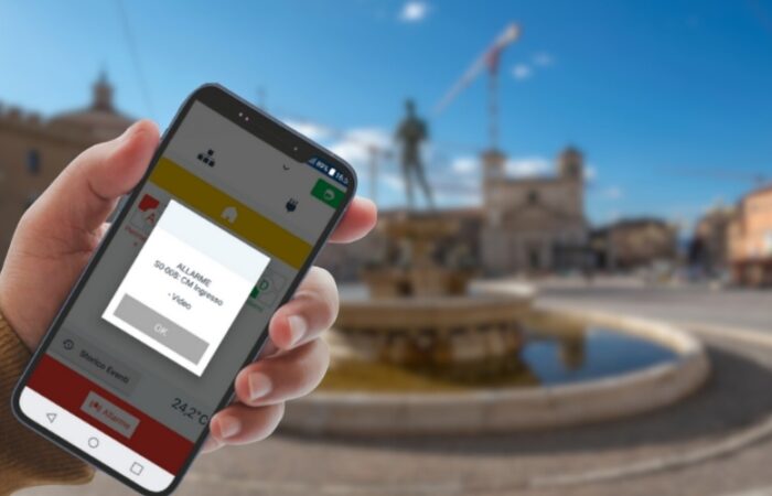 Mano che controlla l’allarme antifurto tramite smartphone davanti alla fontana di Piazza Duomo a L’Aquila.